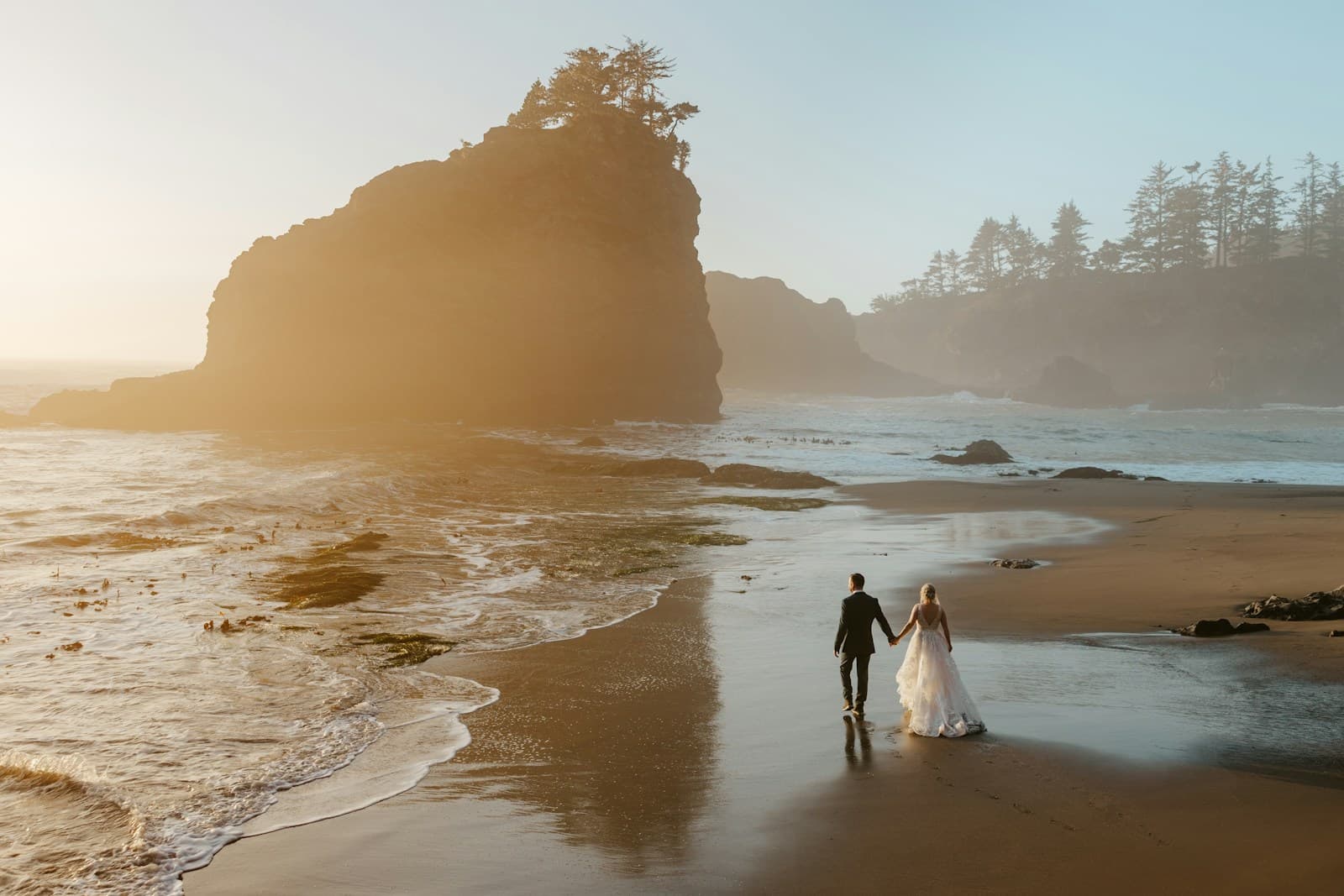 A couple walking along a sunlit beach beneath a rocky headland. Temporary editorial photograph.