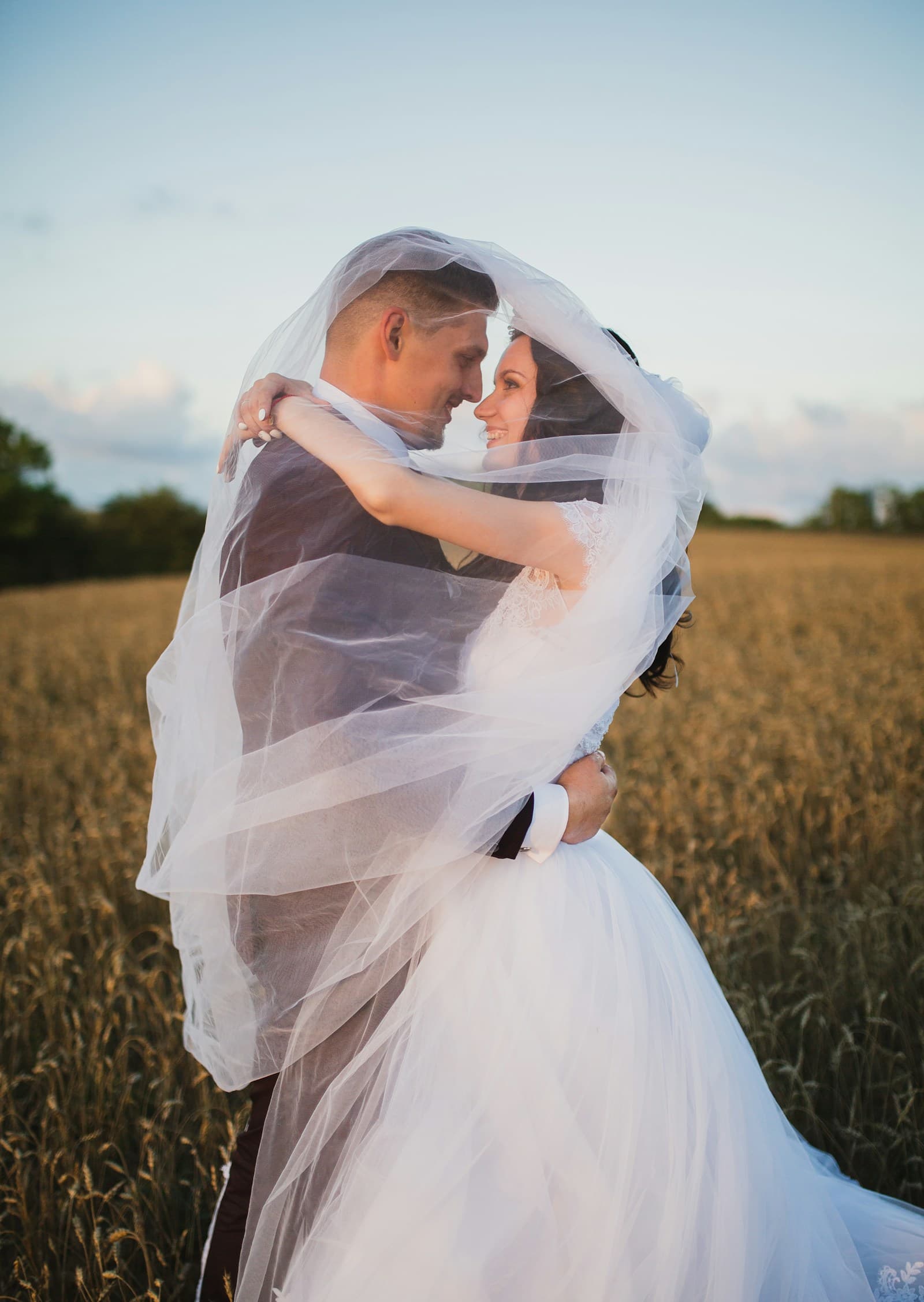 A newlywed couple wrapped in a veil in an open field. Temporary editorial photograph.